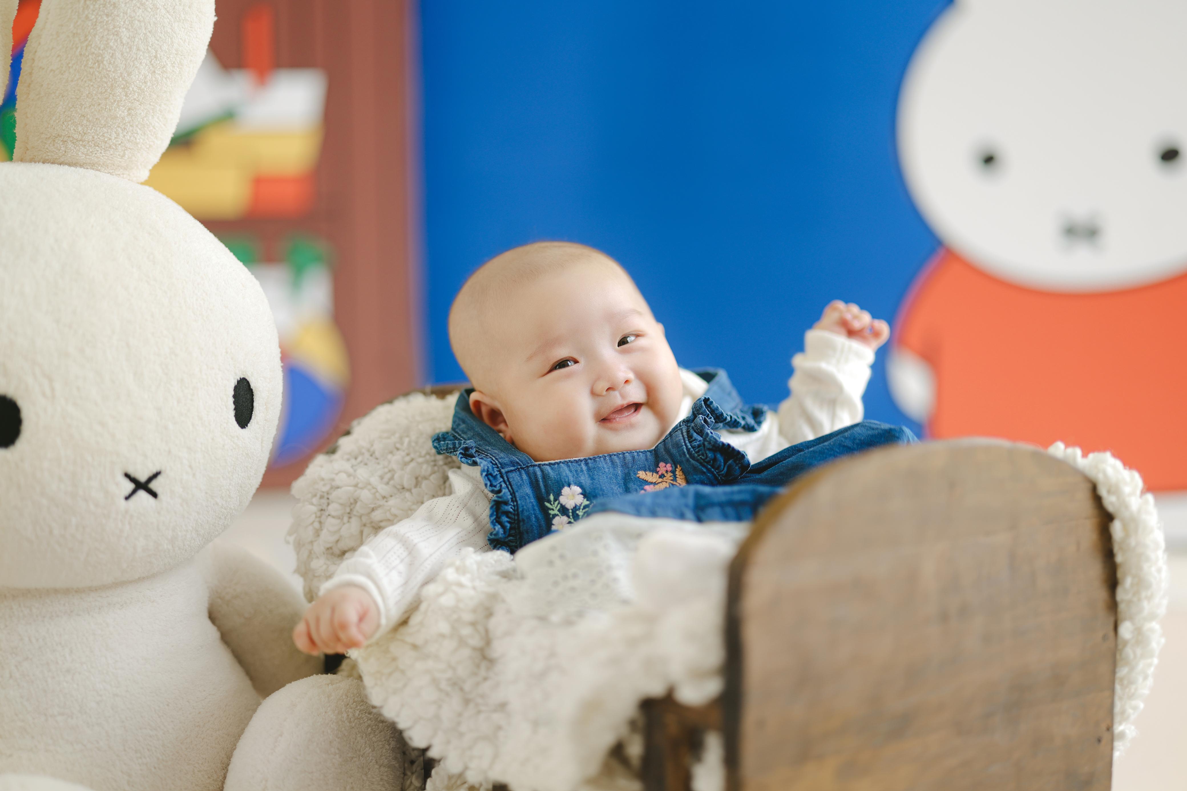 Bayi tersenyum di keranjang kayu dengan mainan lembut, satu tangan terangkat gembira — kehangatan layanan anak terpercaya sejak hari pertama.